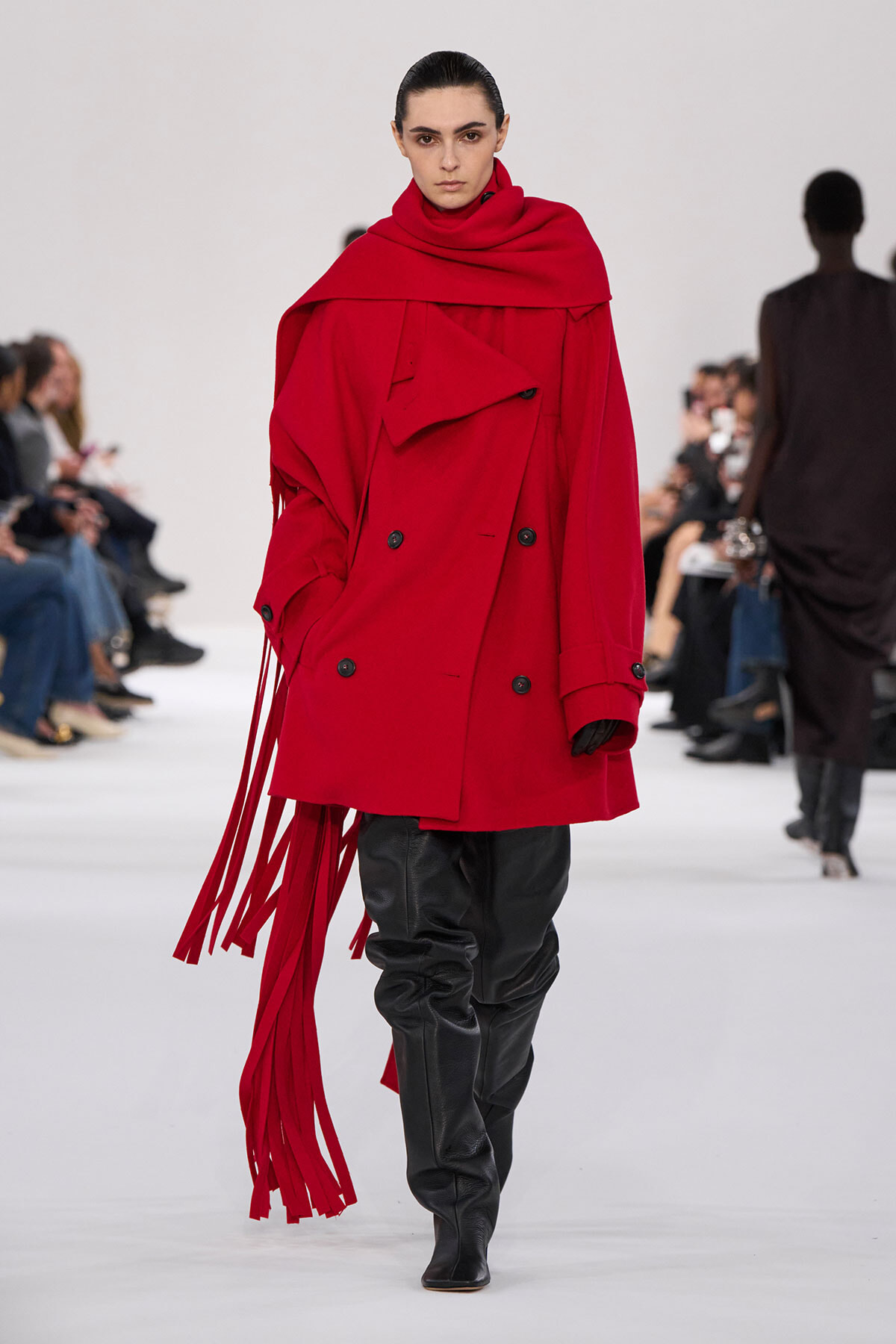 Model walking a runway in a bright red oversized trench with fringe details and black leather pants under black boots.