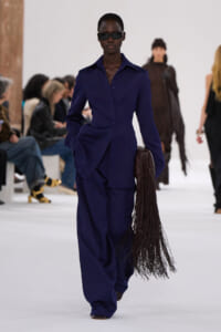 Model walks a fashion runway in a cobalt blue tailored pantsuit, wearing dark sunglasses and carrying a long brown fringe bag at her side.