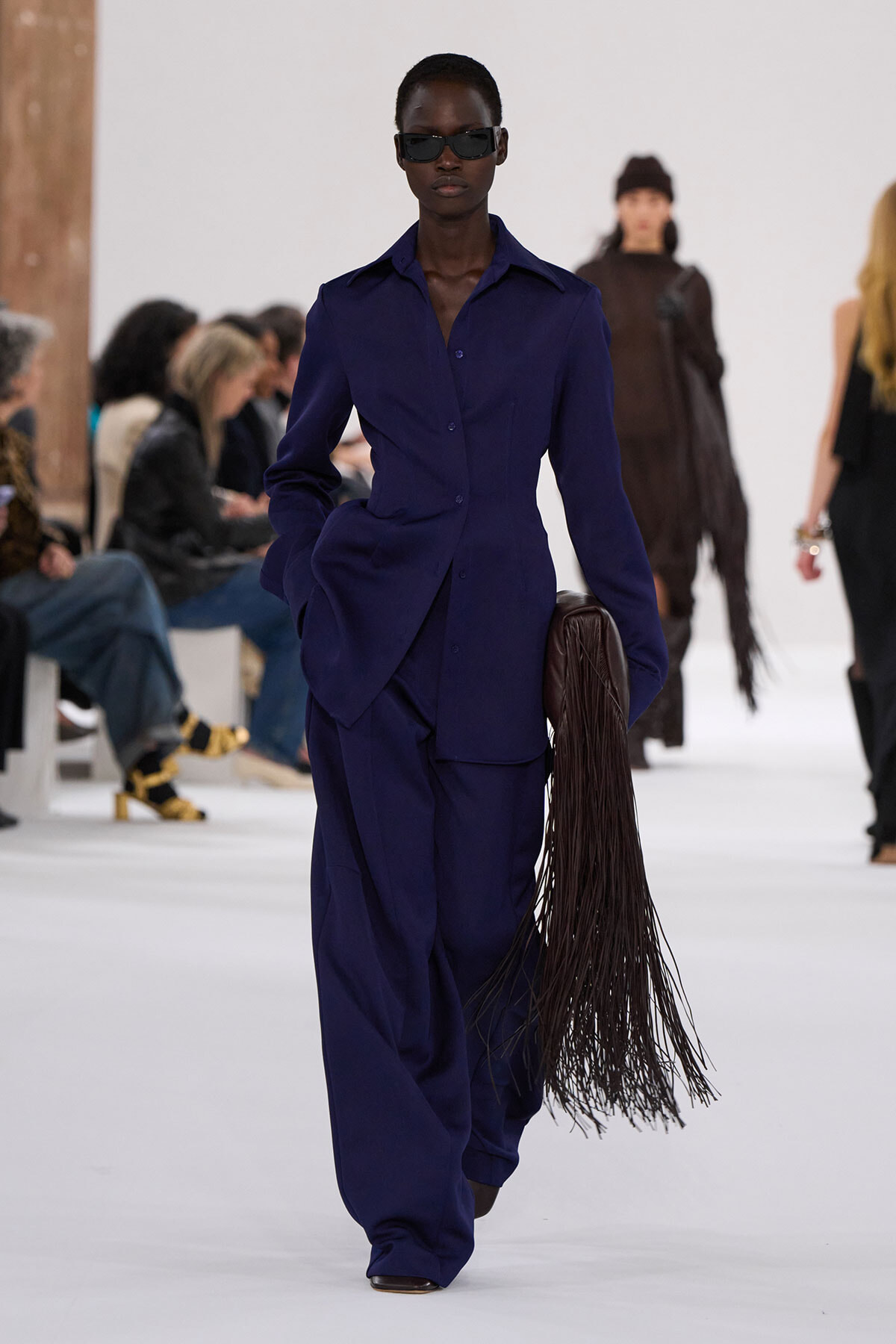 Model walks a fashion runway in a cobalt blue tailored pantsuit, wearing dark sunglasses and carrying a long brown fringe bag at her side.