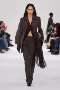 Female model walking down a fashion show runway in a dark plum pantsuit with a long fringe bag.