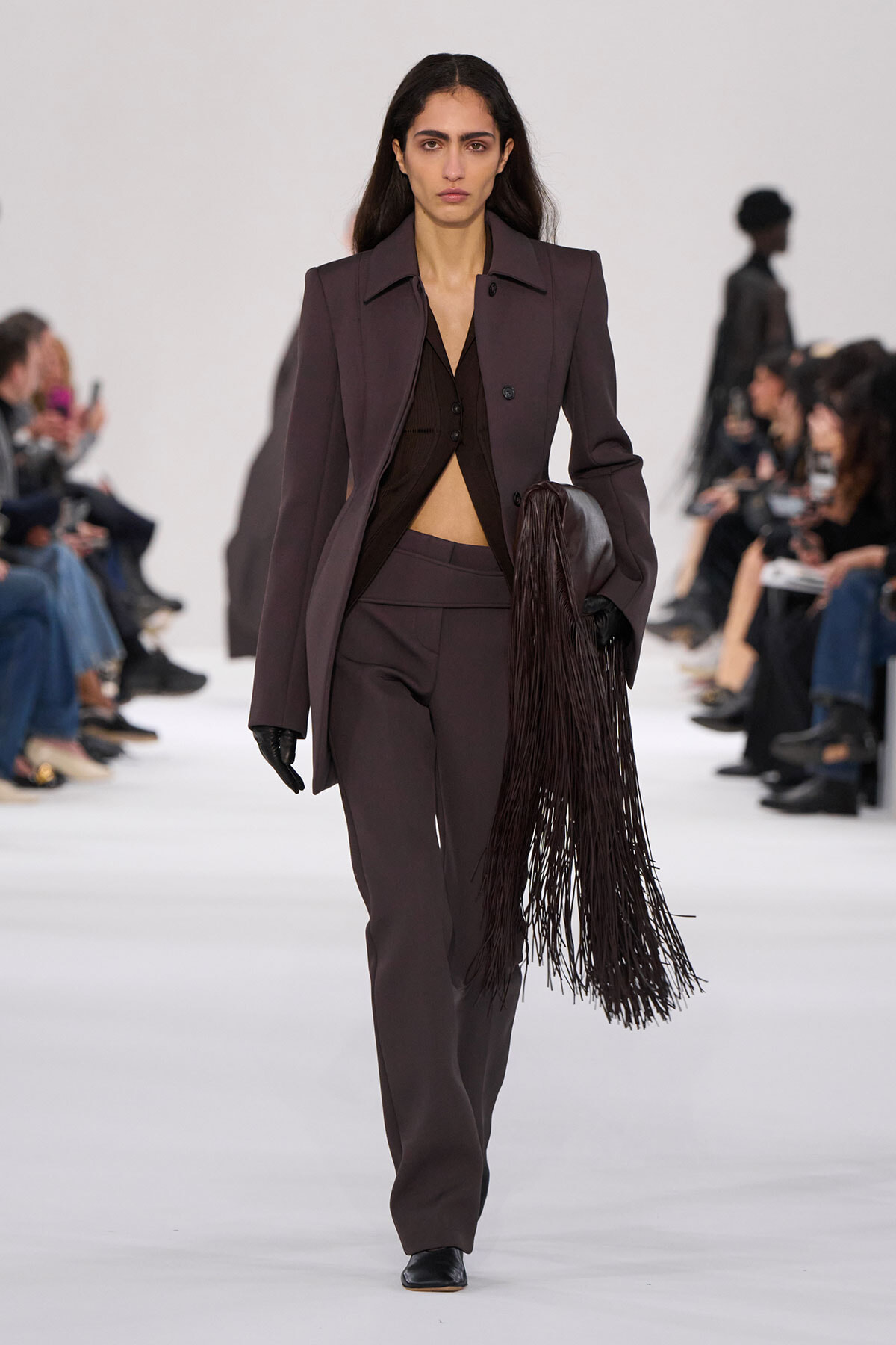 Female model walking down a fashion show runway in a dark plum pantsuit with a long fringe bag.