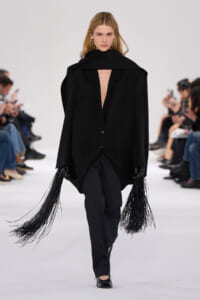 Model walks a minimalist black outfit: oversized blazer with deep V-neck, long fringe tassels from sleeves, black pants, and heels on a runway.