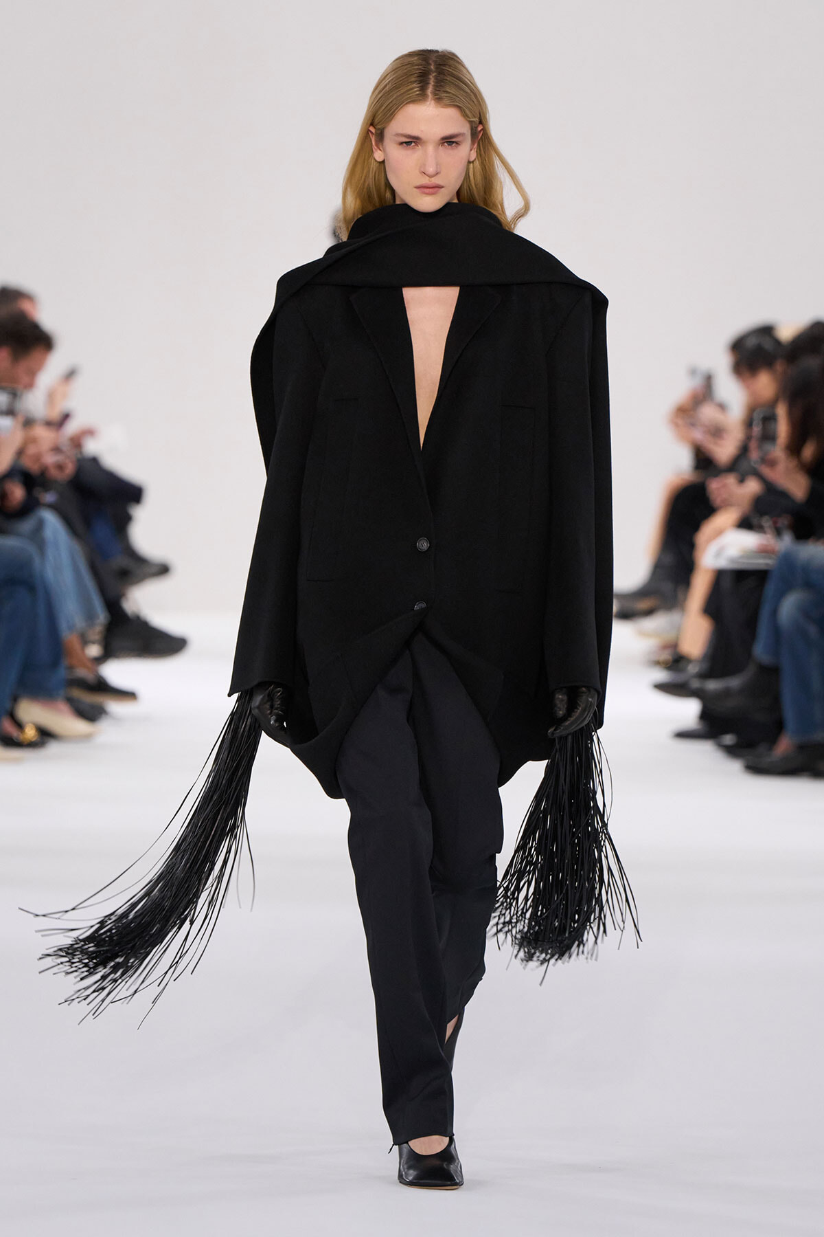 Model walks a minimalist black outfit: oversized blazer with deep V-neck, long fringe tassels from sleeves, black pants, and heels on a runway.