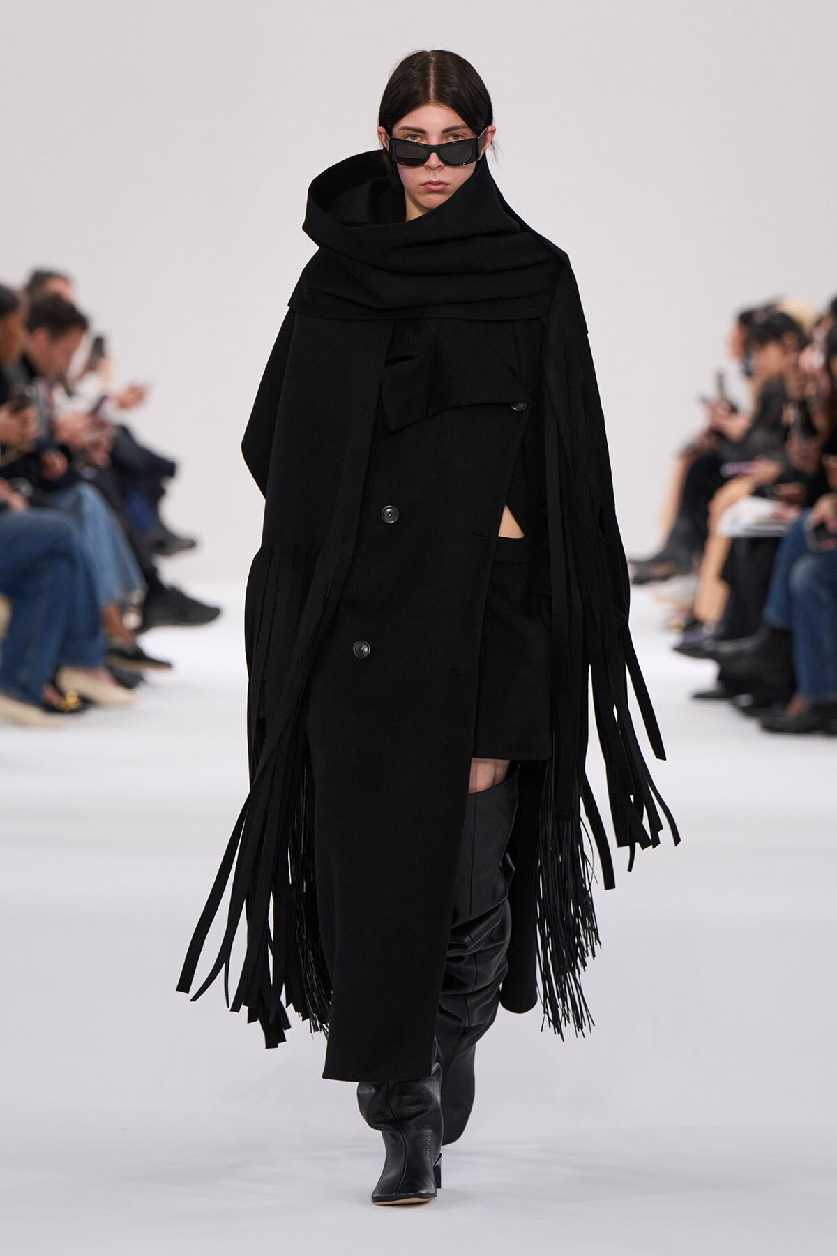 Female model walks a runway in an all-black layered coat with long fringe panels and oversized scarf, wearing black sunglasses and knee-high boots.