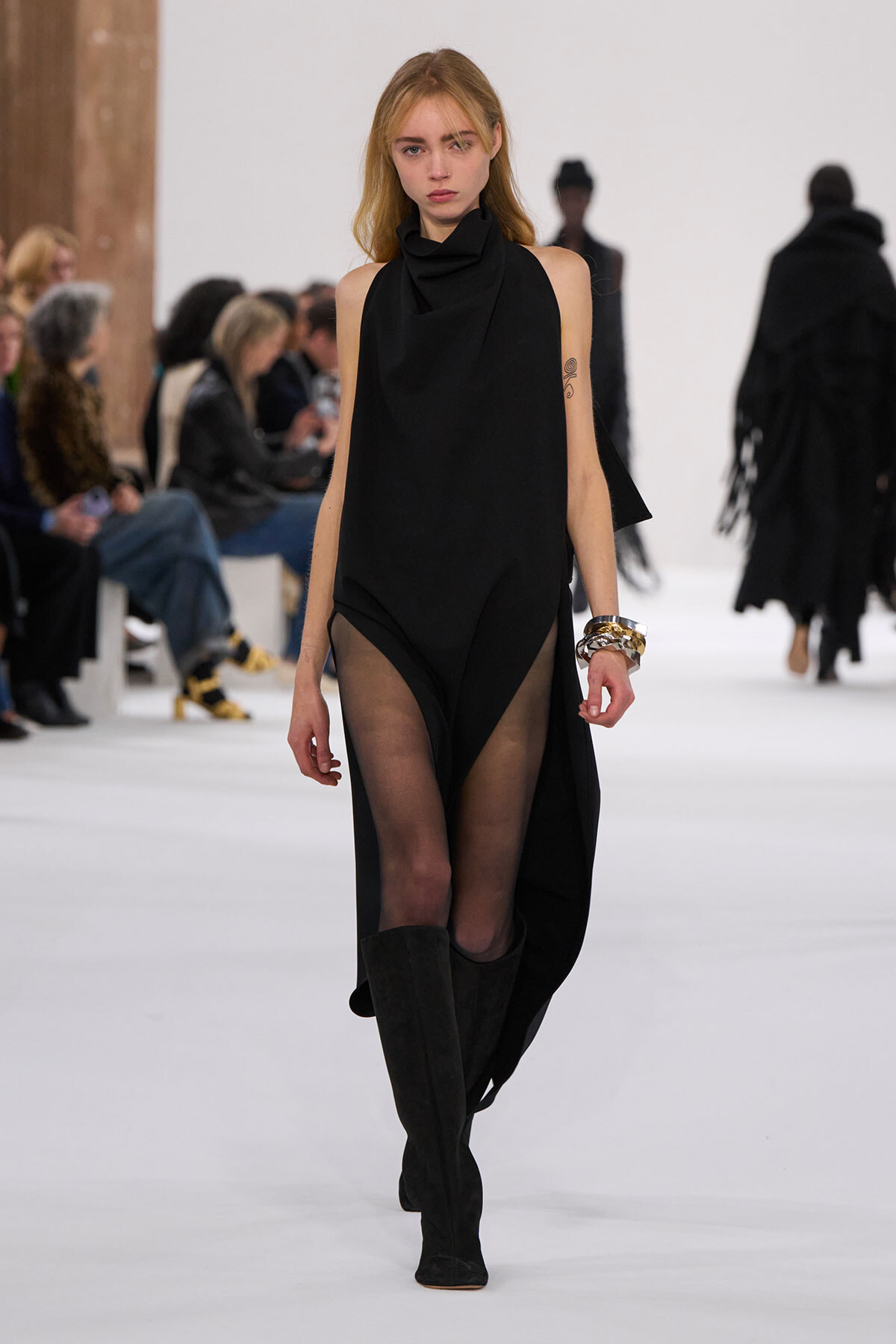 Model walks a fashion runway in a black sleeveless draped dress with sheer tights and knee-high black boots; audience in the background.