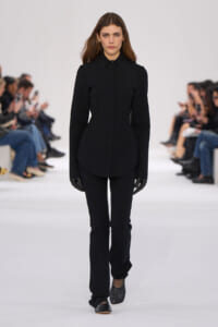 Model walks the runway in an all-black structured outfit: a fitted high-collar coat with curved seams, matching black pants, and gloves.