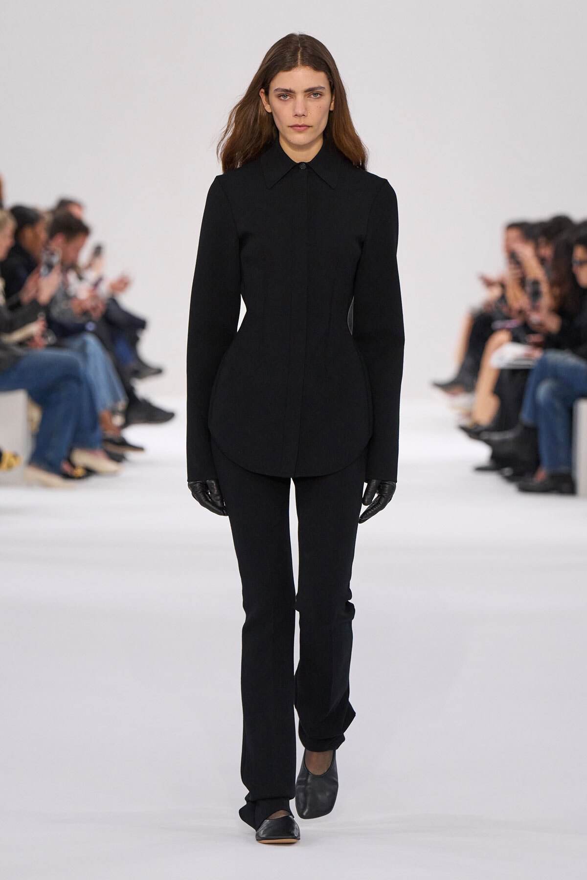 Model walks the runway in an all-black structured outfit: a fitted high-collar coat with curved seams, matching black pants, and gloves.