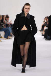 Model walks the runway in an all-black ensemble: long textured coat open at the midriff over a cropped top and black shorts, with sheer tights and black shoes.