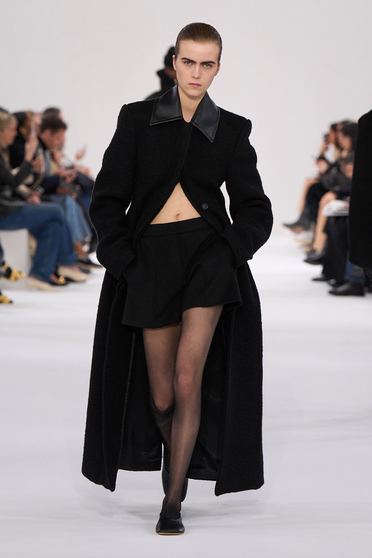 Model walks the runway in an all-black ensemble: long textured coat open at the midriff over a cropped top and black shorts, with sheer tights and black shoes.