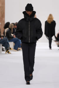 Model walking a runway in an all-black outfit: oversized fur-curred zip jacket, black pants, and a black beanie.