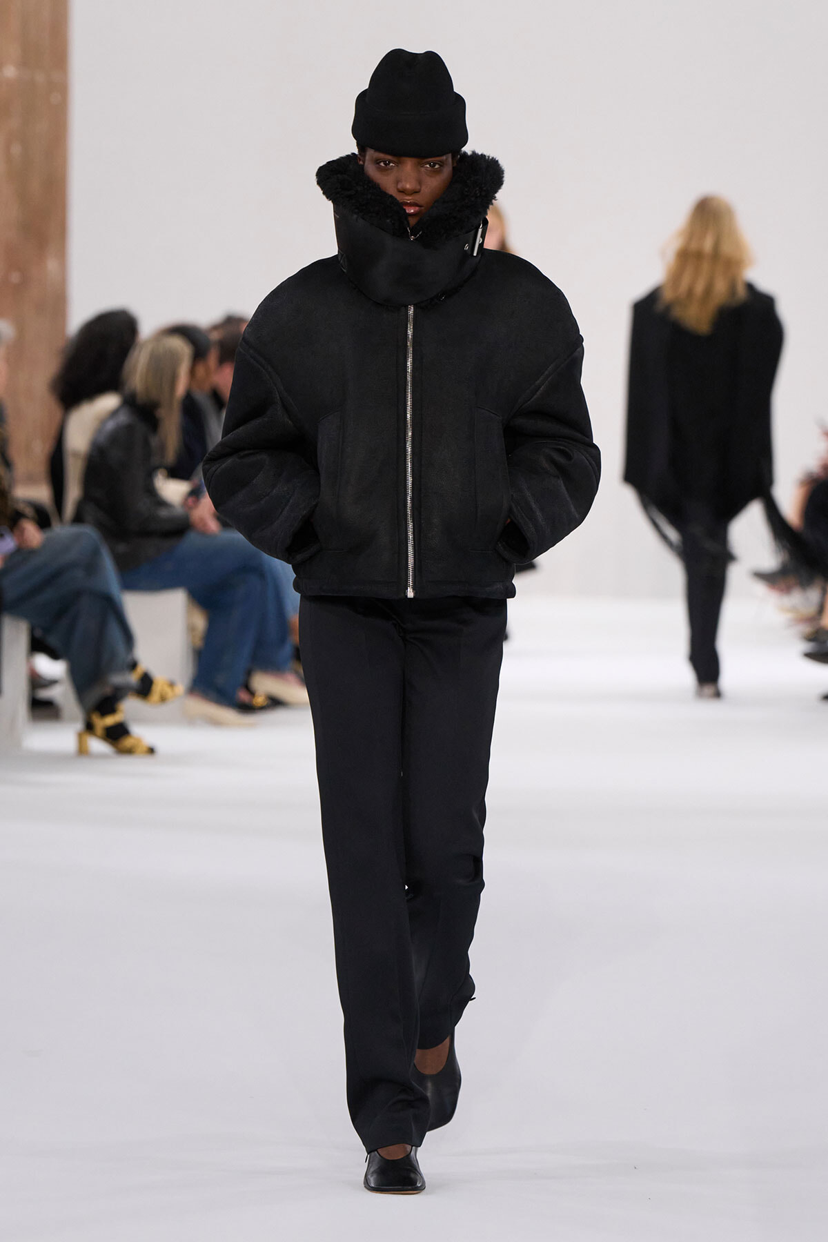 Model walking a runway in an all-black outfit: oversized fur-curred zip jacket, black pants, and a black beanie.