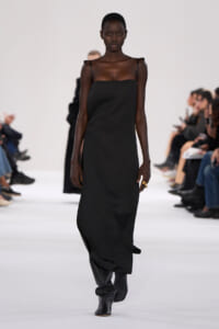Model walks the runway in a black spaghetti-strap slip dress and tall black boots, audience out of focus in the background.