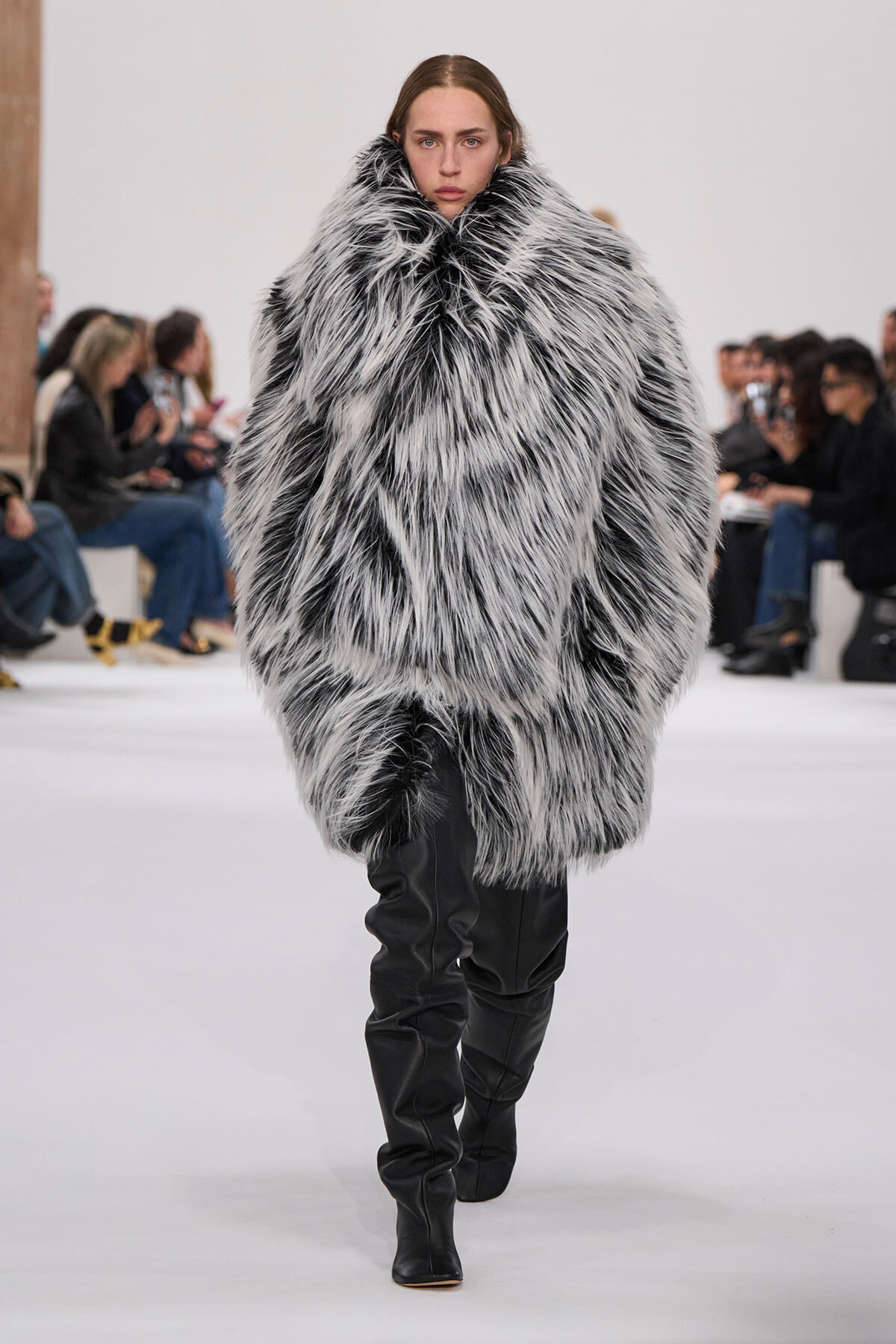 Model walking a fashion runway in a oversized black-and-white faux fur coat with knee-high black boots.