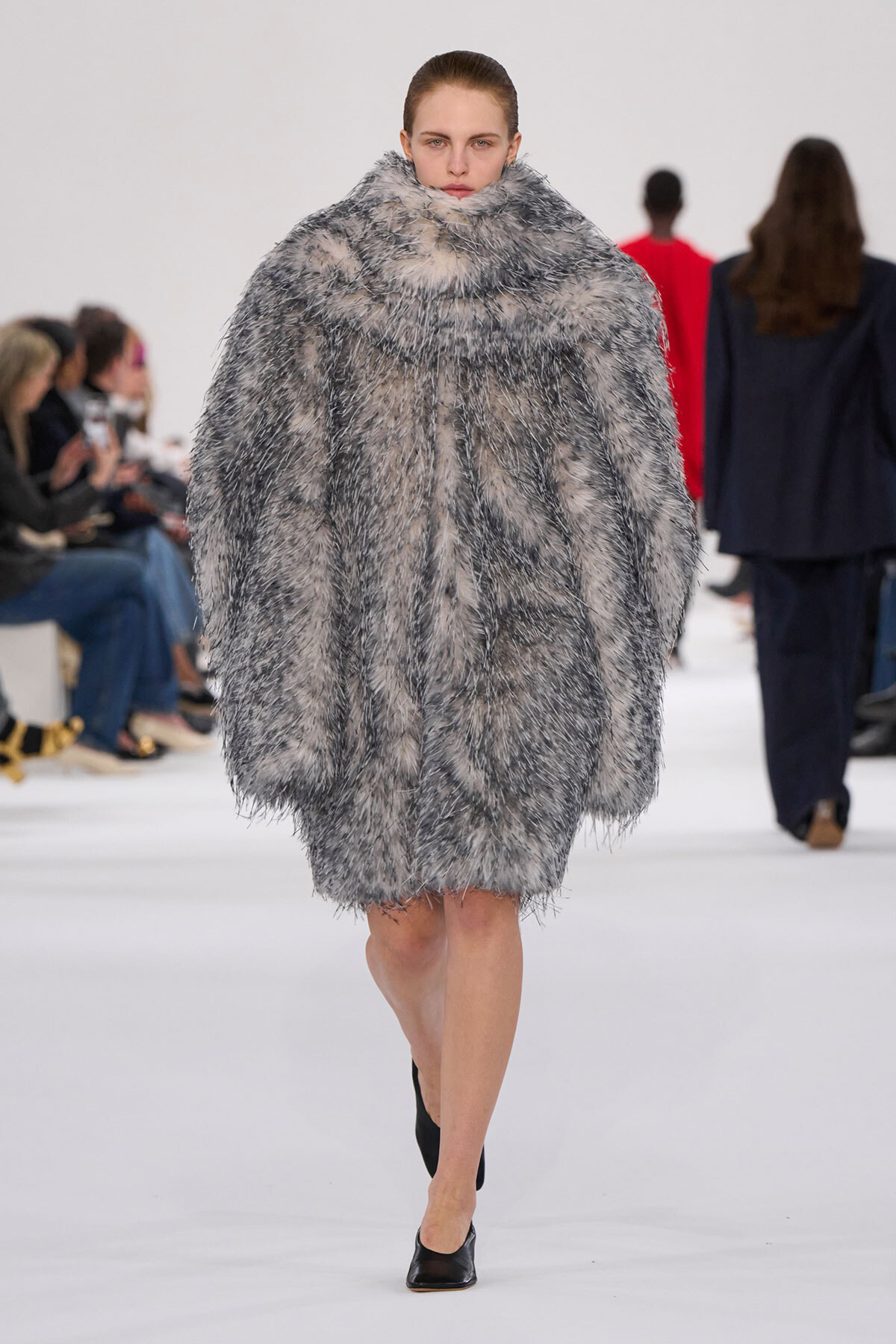 Model on a runway wearing an oversized gray shaggy fur coat, with spectators seated along the sides in the background.