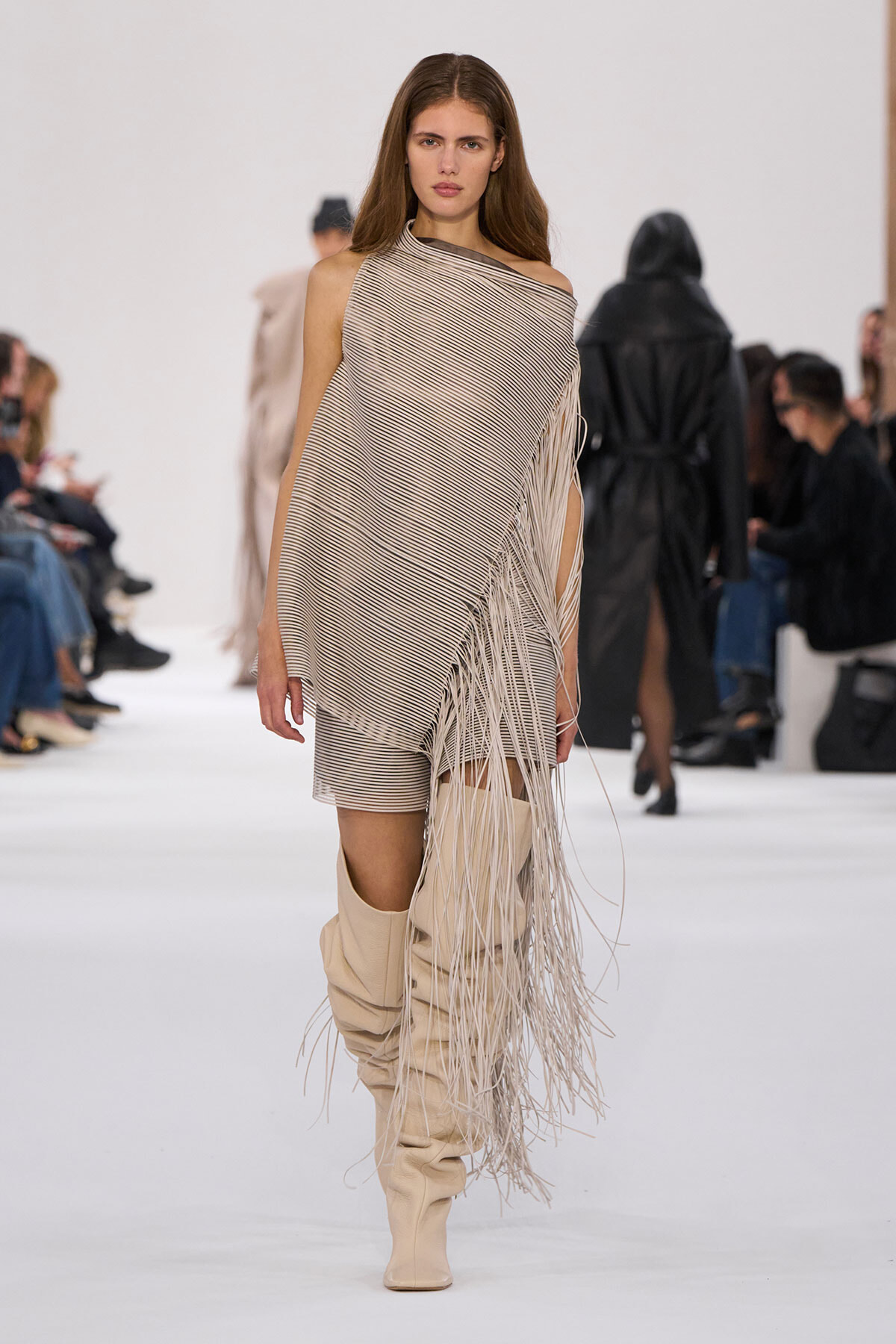 Model walks the runway in a beige and black diagonal striped one-shoulder poncho with long fringe and beige knee-high boots.