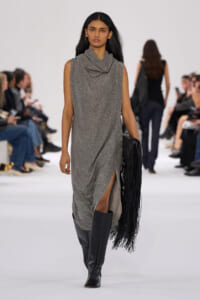 Model walking a runway in a glittery gray sleeveless dress with a draped collar, knee-high black boots, and a fringed black handbag.