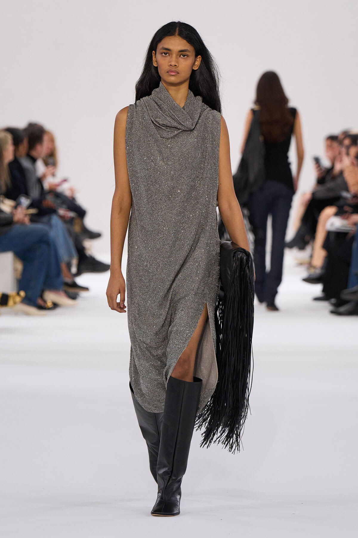 Model walking a runway in a glittery gray sleeveless dress with a draped collar, knee-high black boots, and a fringed black handbag.