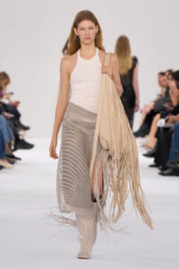 Model walking a fashion show wearing a white sleeveless top, striped beige skirt, knee-high cream boots, and a large beige fringe bag over her shoulder.