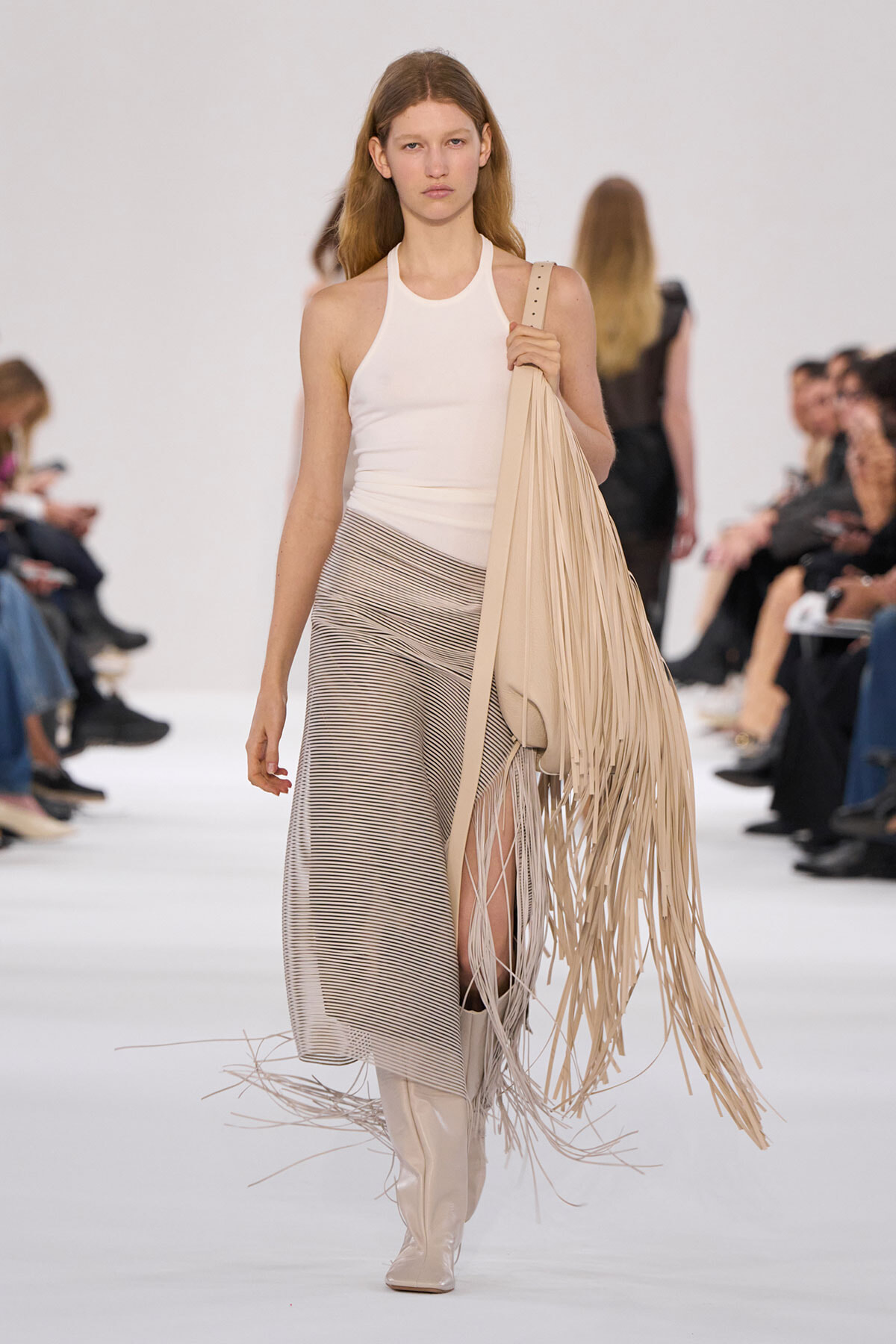 Model walking a fashion show wearing a white sleeveless top, striped beige skirt, knee-high cream boots, and a large beige fringe bag over her shoulder.