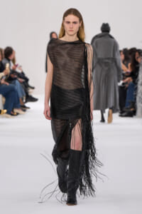 Model walks a fashion show in a sheer black striped dress with long fringe, paired with knee-high black boots.