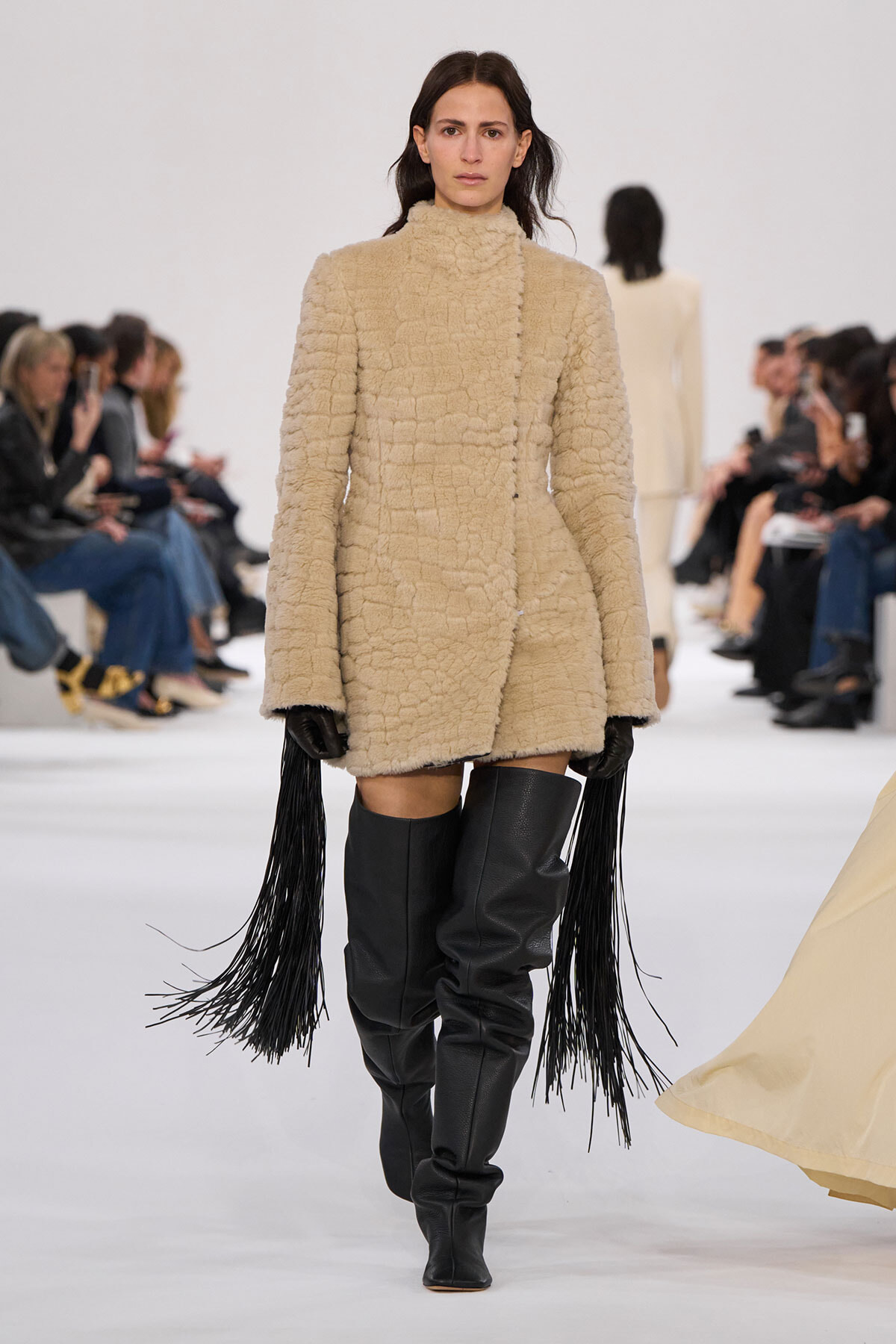 Model walks the runway in a beige textured coat with black thigh-high boots; audience seated along the sides watch.