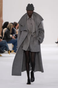 Model walking a runway in a monochrome gray ensemble: oversized ribbed coat, matching shirt and shorts, black tights and a gray hat.