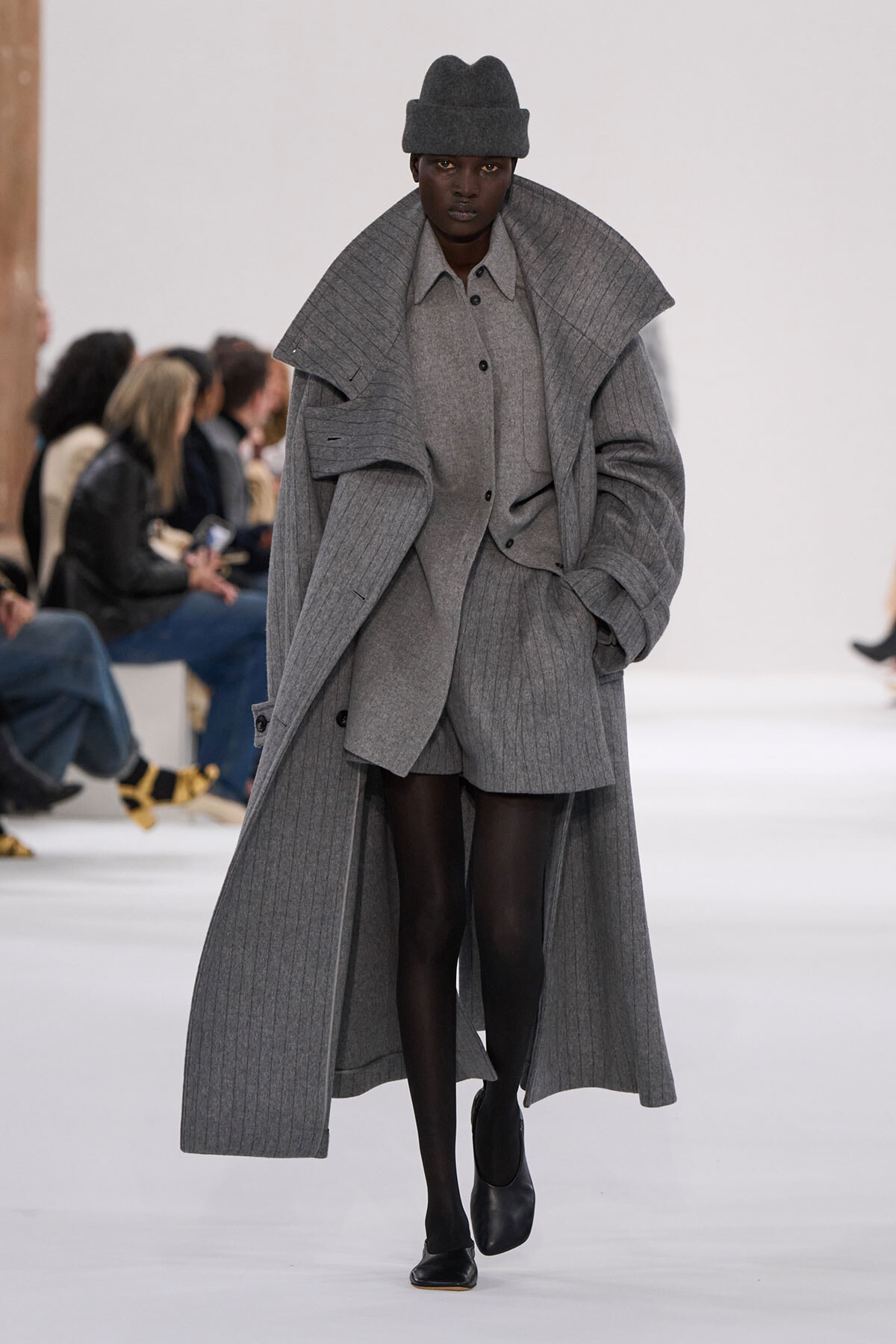 Model walking a runway in a monochrome gray ensemble: oversized ribbed coat, matching shirt and shorts, black tights and a gray hat.