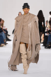 Model walks the runway in a taupe fringe cape over a matching outfit with knee-high beige boots and a black leather hat on a white set stage.