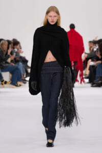 Model walks a fashion show wearing a black asymmetrical sweater exposing midriff, dark jeans, black gloves, and a long fringe bag on the right.