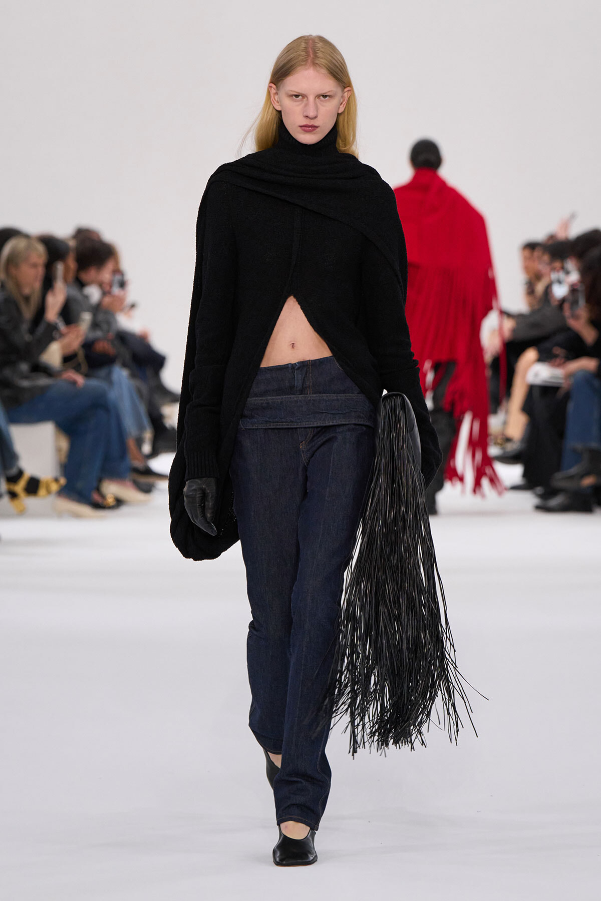Model walks a fashion show wearing a black asymmetrical sweater exposing midriff, dark jeans, black gloves, and a long fringe bag on the right.