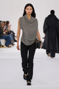 Model walking a runway in a glittery gray draped sleeveless top and black pants, carrying a black clutch accessory.