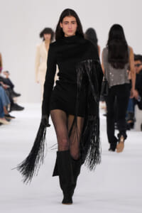 Model walks a fashion show in a black mini dress with long fringe draping from the bodice and sleeves, paired with knee-high black boots.