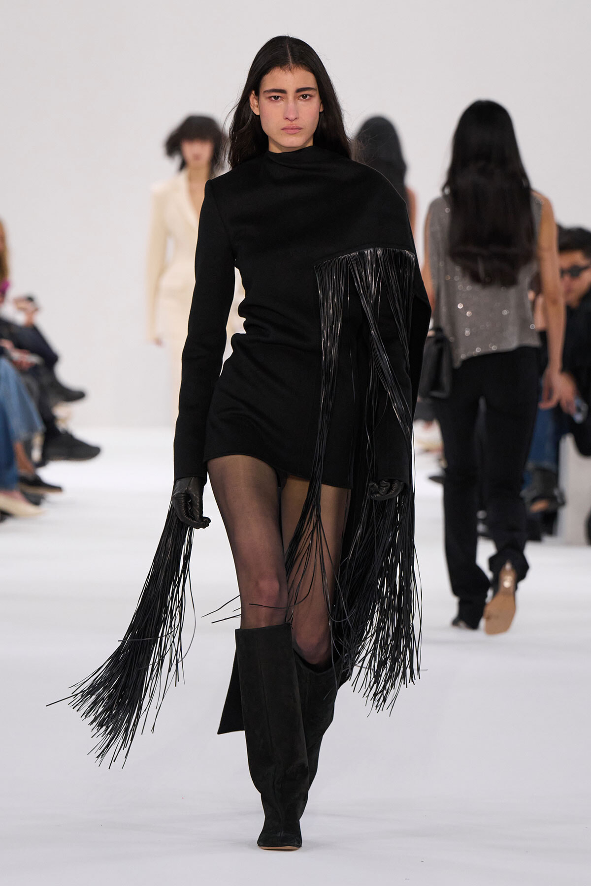 Model walks a fashion show in a black mini dress with long fringe draping from the bodice and sleeves, paired with knee-high black boots.