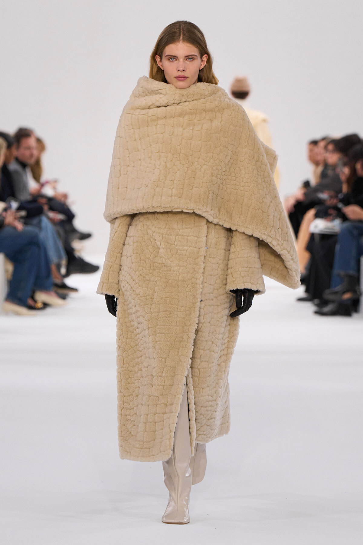 Model on a fashion runway wearing an oversized beige shearling cape over a matching coat and beige knee-high boots; audience sits along the sides in the background.