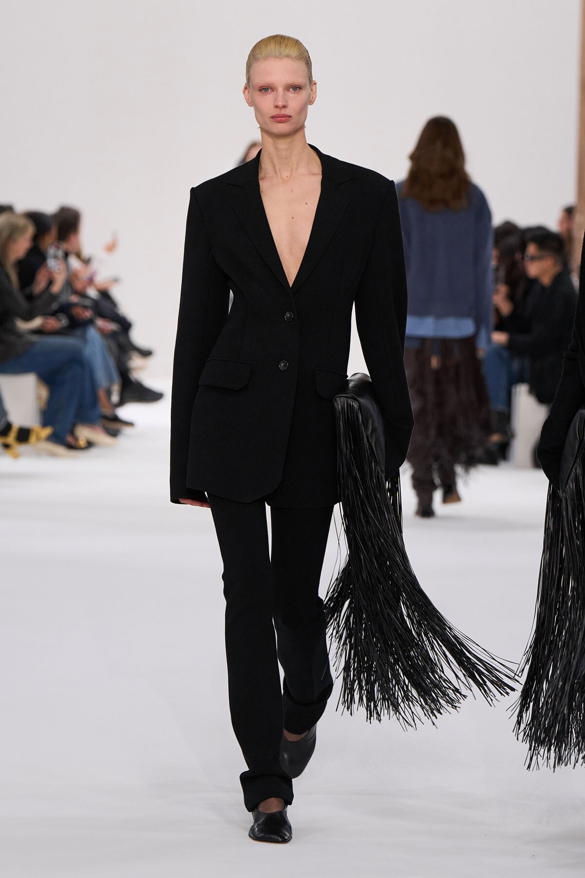 Model on a white runway wearing a black tailored blazer and slim pants with a deep V neckline; long black fringe accessories trail from her right side, as onlookers sit along the sides of the catwalk.