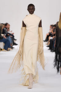Model on a runway wearing a cream sleeveless turtleneck dress with long fringe sleeves and matching fringe cape.