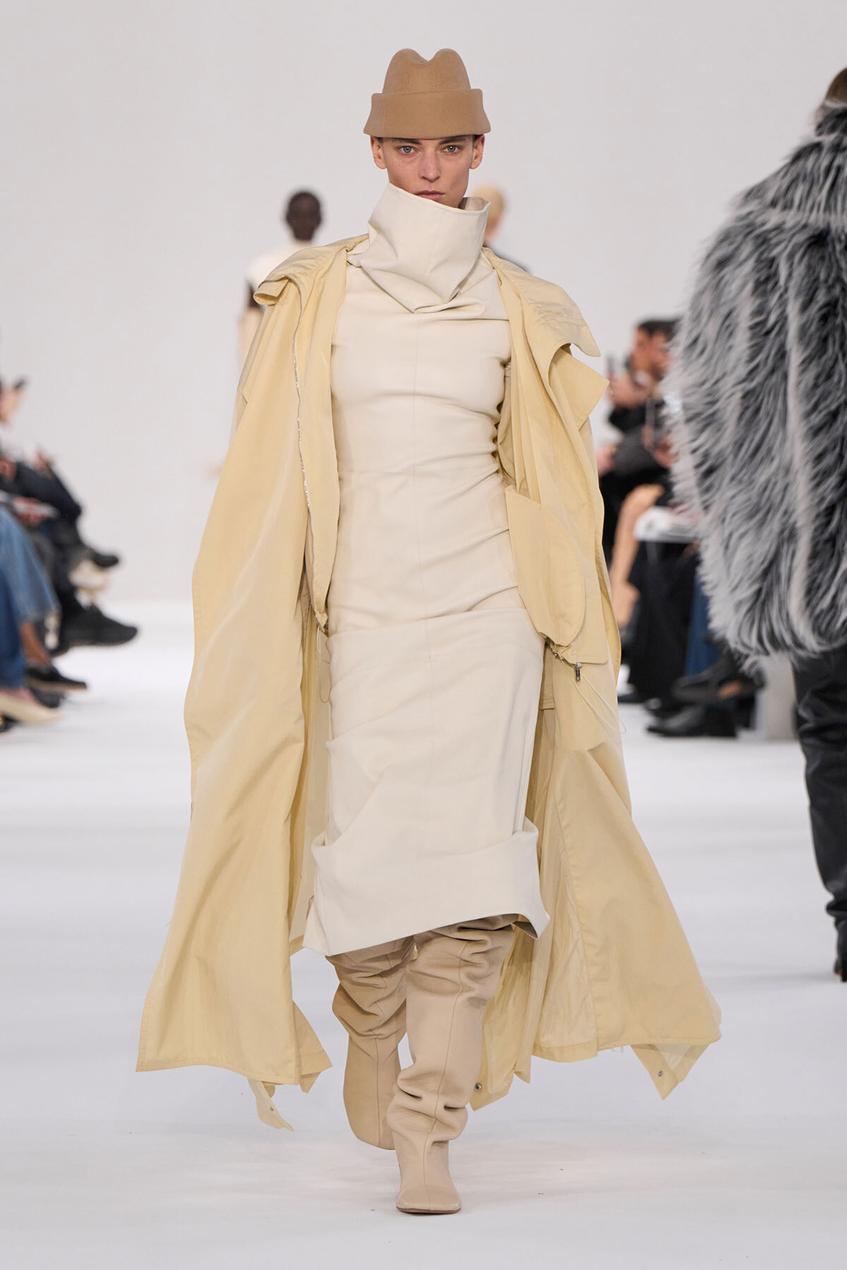 Model walks the runway in a monochrome beige ensemble: high-collared sheath dress, oversized trench draped over shoulders, and tan knee-high boots with a matching fedora.