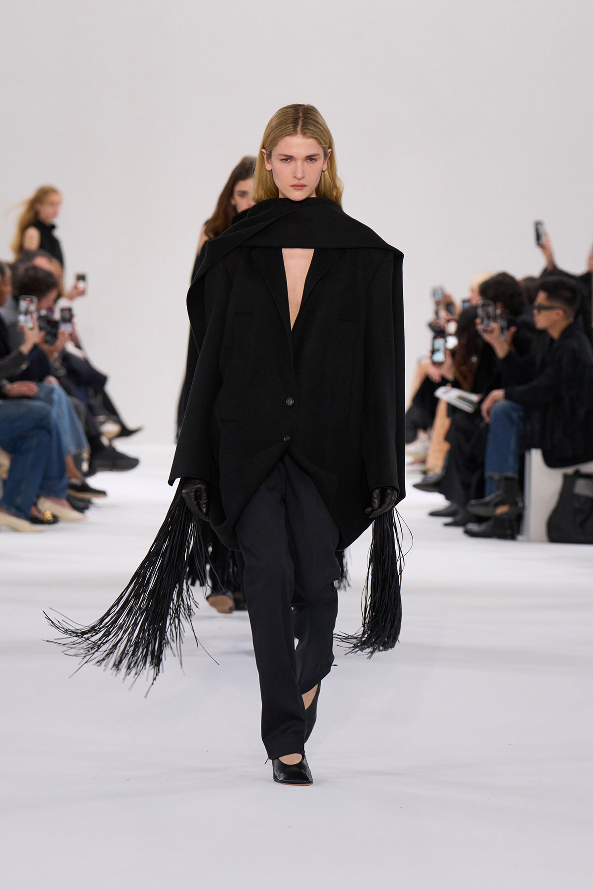 Model on a runway wearing an oversized black coat with a cape-like collar and fringe panels, walking toward the camera. The V-neck opening is visible beneath the coat edge.