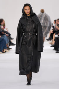 Model on a fashion runway wearing a long black leather hooded coat with a waist belt.
