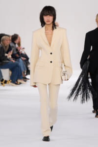 Model on a fashion runway wearing a cream tailored blazer and matching trousers, carrying a small cream handbag.