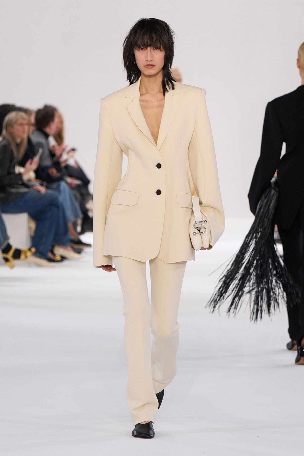 Model on a fashion runway wearing a cream tailored blazer and matching trousers, carrying a small cream handbag.