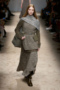 Model walking a runway in a textured gray knit outfit with a draped cape-like overlay and matching belt, carrying a gray shoulder bag and wearing brown boots.