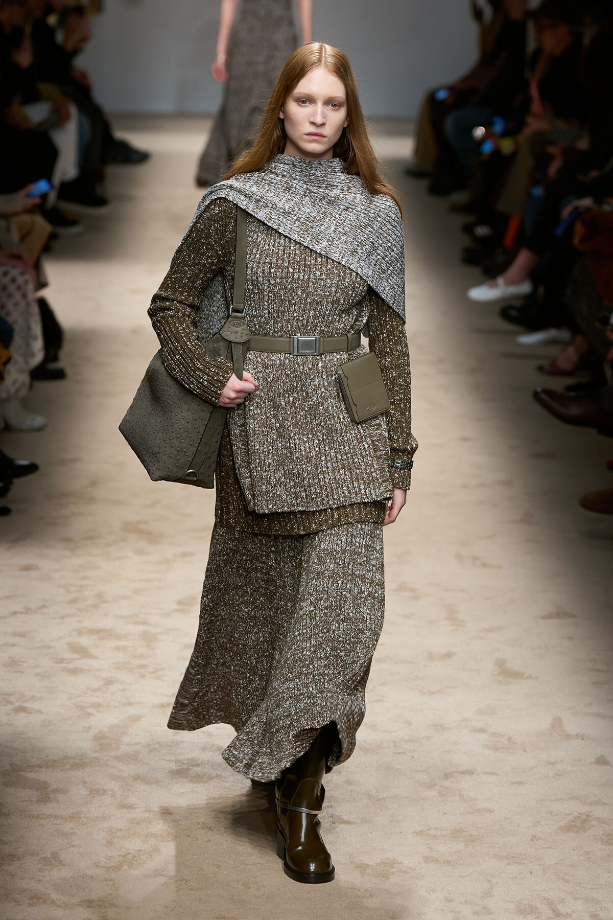 Model walking a runway in a textured gray knit outfit with a draped cape-like overlay and matching belt, carrying a gray shoulder bag and wearing brown boots.