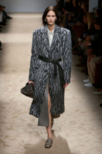 Runway model wearing a long grey fur coat belted at the waist, carrying a brown quilted handbag.