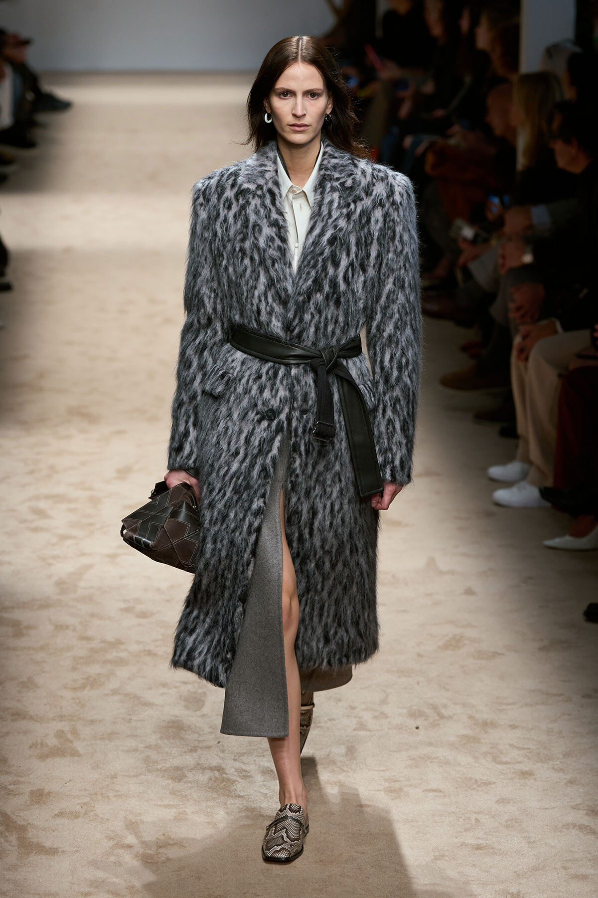 Runway model wearing a long grey fur coat belted at the waist, carrying a brown quilted handbag.