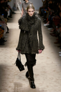 Model walks the runway in a chunky, textured gray coat with fringe, paired with black boots and a black handbag.