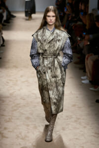 Model walks down a runway wearing a textured gray-brown fur-like trench with a belt and blue-gray sleeves, snakeskin boots visible.