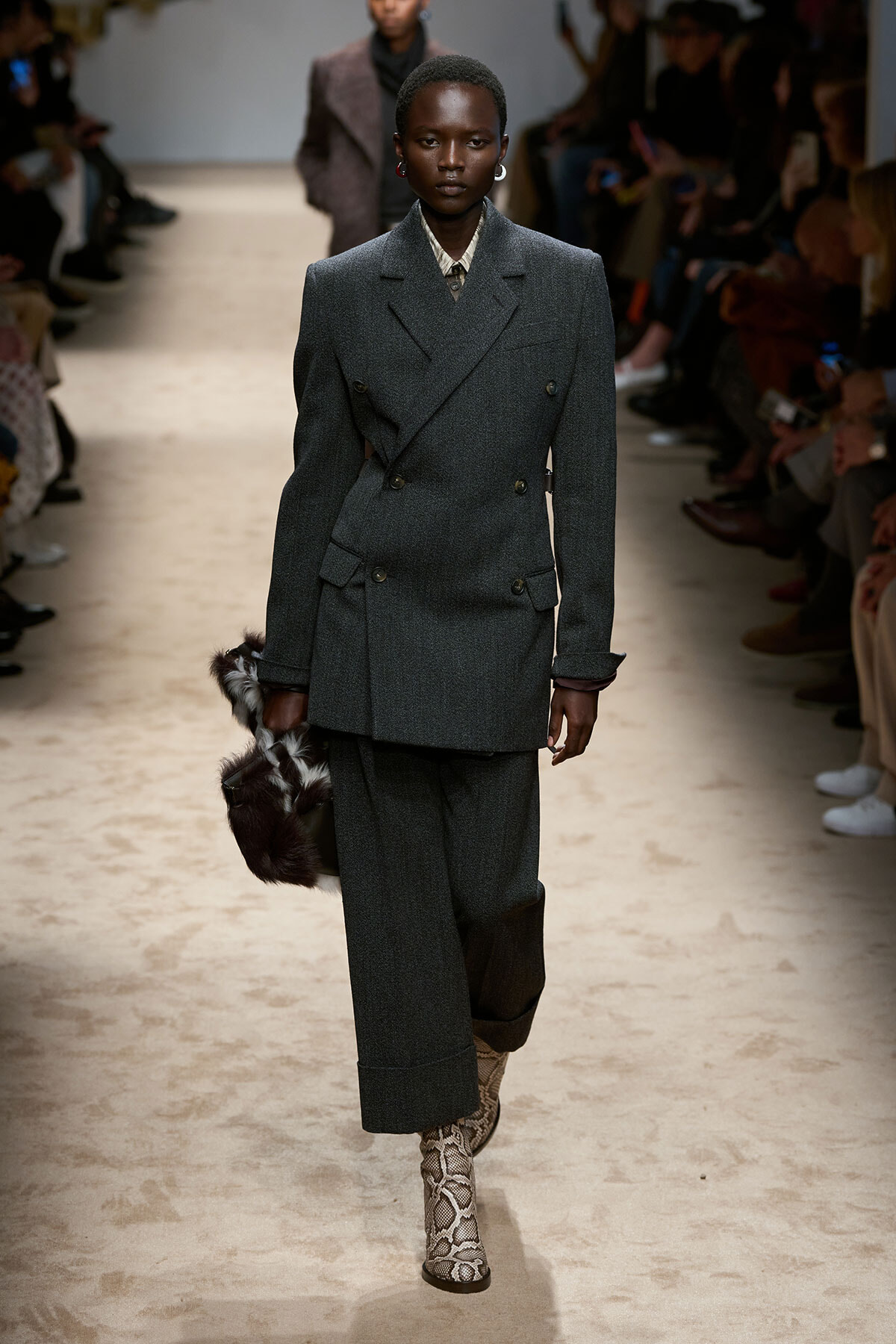 Model on a runway wearing a charcoal gray double-breasted suit with oversized shoulders, holding a fur clutch, and snakeskin boots.