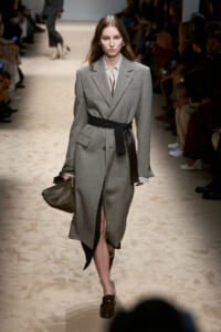 Model walks the runway in a gray tailored wool coat cinched with a black belt, carrying a textured taupe handbag.