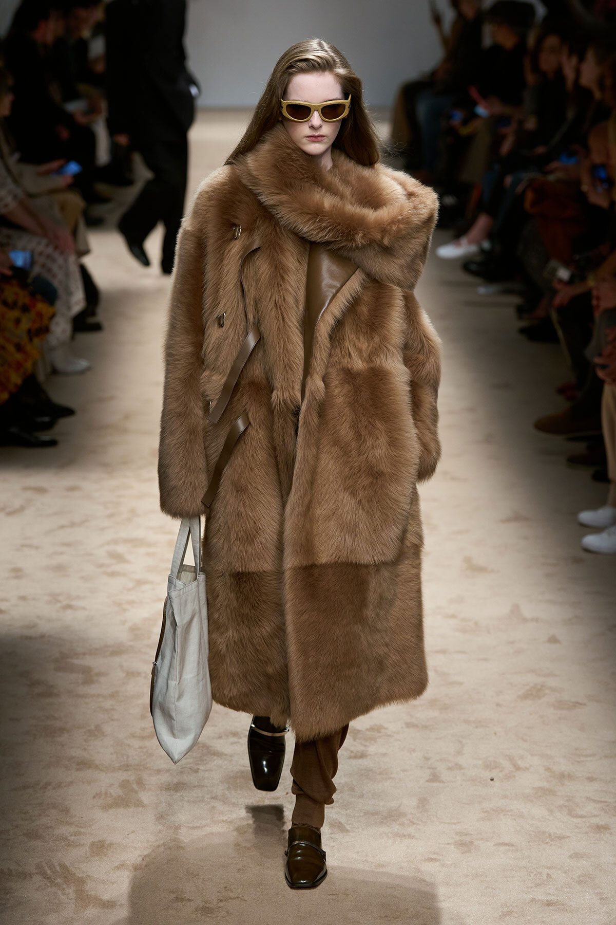 Model walks the runway in a full-length brown fur coat with oversized collar, beige sunglasses, and a light gray tote bag.